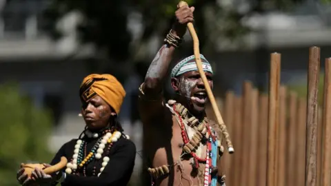 Reuters Two praise singers are seen dressed in colourful traditional attire at a ceremony commemorating the military workers. The praise poet in front holds a cane in one hand.