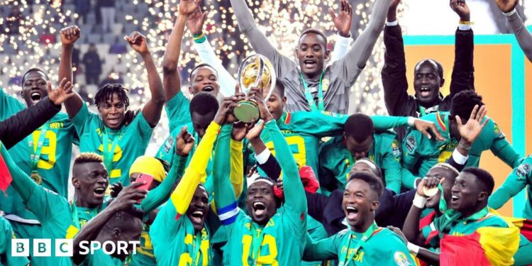 A group of Senegal players wearing green lift the African Nations Championship trophy, with flashes of fireworks seen in the background inside a stadium