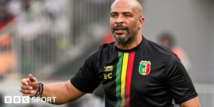 Eric Chelle wears a predominantly black Mali training top, with green, yellow and red stripes on the front and his initials on it as he stands on the touchline during a football match