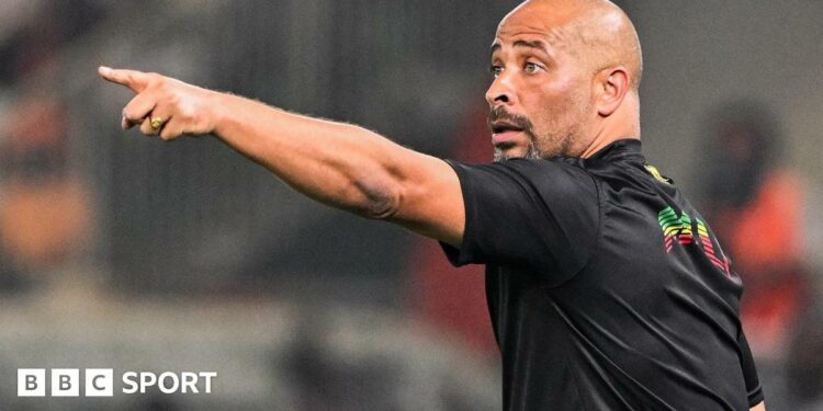 Eric Chelle, wearing a black T-shirt and with his back half turned to camera, points his left arm out in front of him as he gestures during a football match