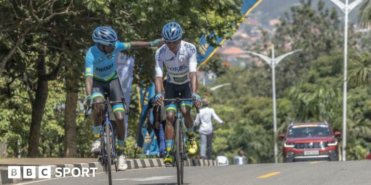 Two cyclists, one wearing a blue jersey and black shorts and the other in a white jersey and black shorts ride along a tree-lined street in Kigali while a maroon car is seen in the background