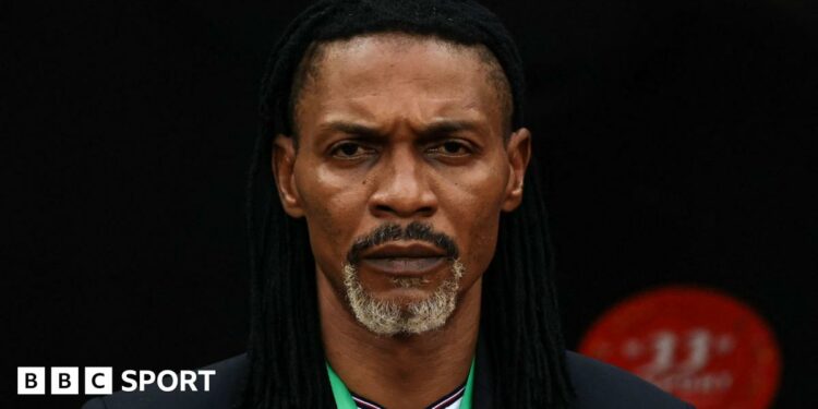 A close-up headshot of Rigobert Song looking sternly into the camera while wearing a black jacket, white shirt and with green lanyard around his neck