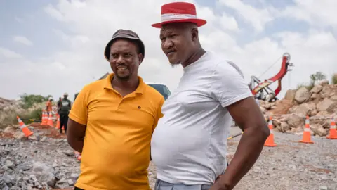 BBC Mandla Charles and Mzwandile Mkwayi, the two community volunteers who descended into the abandoned mine, stand side by side near the top of the mine shaft. The one on the left is wearing a yellow T-shirt and the other is wearing a white T-shirt.