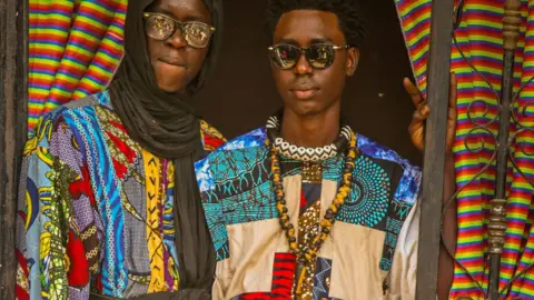 Getty Images Two men in multi-patterned colourful patchwork clothes look at the camera framed by a doorway. One is wearing a black headscarf and glasses, the other is in sunglasses and has beaded necklaces around his neck - in Diourbel, Senegal, April 2024