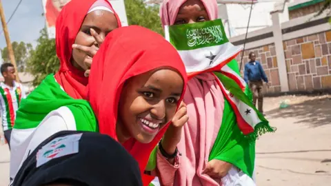 AFP Three young women smiling wearing red and pink headscarves and holding a Somaliland flag and other regalia - Hargeisa, 2018