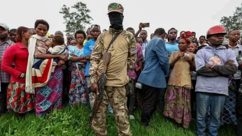 EPA A member of the M23 in a balaclava and with an automatic weapons stands in front of a crowd of civilians.