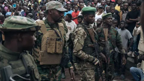 AFP Armed M23 fighters in military uniform and green berets stand in front of a crowd of people. 