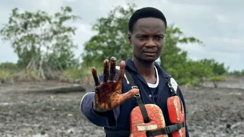 A Nigerian fisherman wearing a navy shirt and a battered orange foam life jacket stands amid an oil-contaminated landscape in Ogoniland. He holds up his hand to the camera - showing his hand stained with oil that is present in the water.