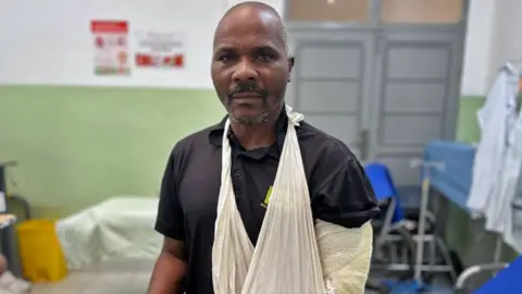 BBC A man in a black t-shirt and jeans sits on a hospital bed. He wears a sling and a bandage around his arm.