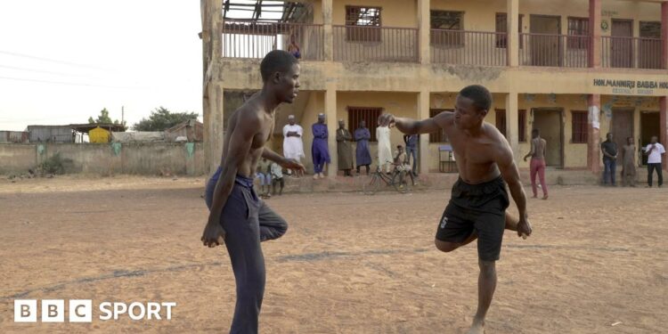 Two topless men are seen opposite each other while balancing on one foot on a dusty patch of land, with several onlookers standing or sitting in front of a two-storey building in the distance